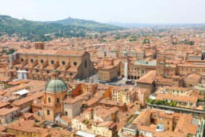 Aerial view of Bologna, Italy, unveils the city’s red-tiled roofs and historic architecture. Prominent is the Basilica of San Petronio with its unfinished façade. This stunning cityscape, ideal for an Italy road trip, is framed by green hills under a clear sky.